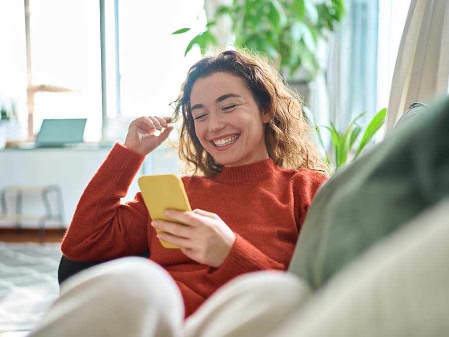 Happy,Relaxed,Young,Woman,Sitting,On,Couch,Using,Cell,Phone,